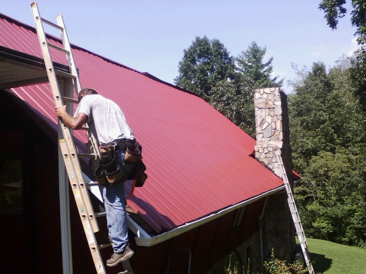 Roofer on a ladder beside a red metal roof and stone chimney — Soffit & Fascia Repair by Roofing Brothers in Savonburg